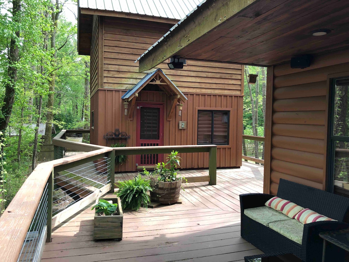 A wooden deck wraps around the main cabin, featuring potted plants and a seating area with cushions. The entrance to the cabin is highlighted by rich wooden siding and a welcoming door, framed by lush greenery that creates a serene atmosphere.