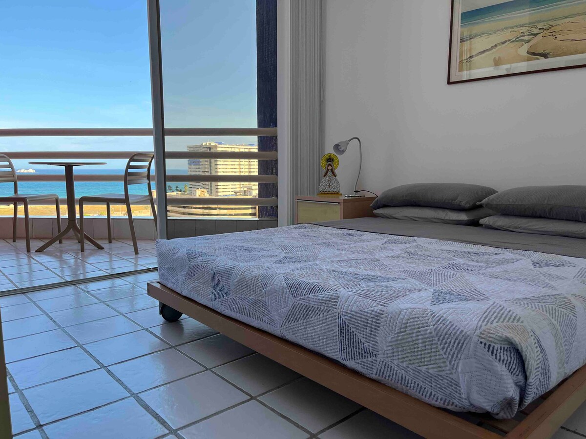 A spacious bedroom features a low-lying bed draped with a patterned quilt. Natural light floods the room, highlighting a small dining table and chairs positioned near a balcony that overlooks a view of the ocean and distant beach.