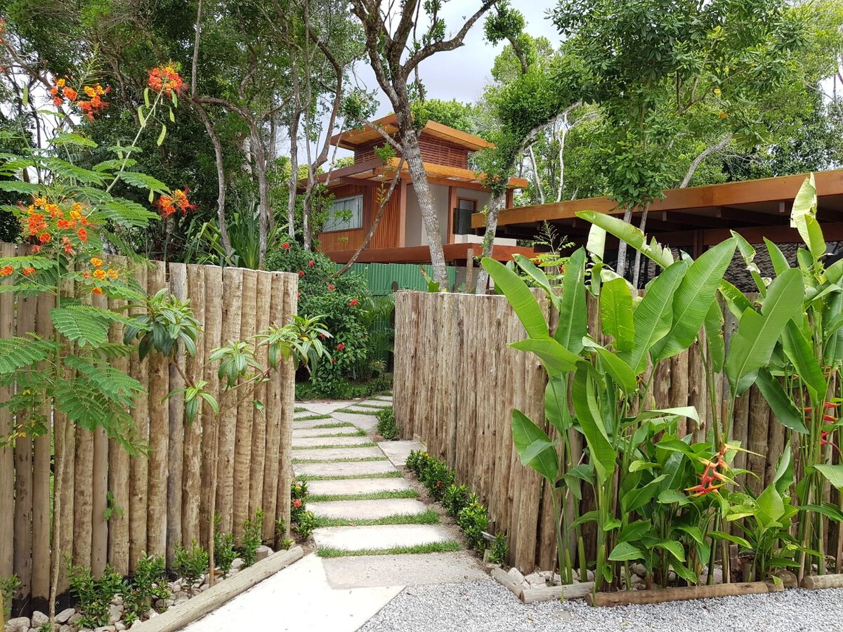 A pathway lined with stepping stones is framed by vibrant greenery and wooden fencing. Tropical plants and flowers add color to the scene, while a glimpse of the charming house is visible in the background, surrounded by lush trees.
