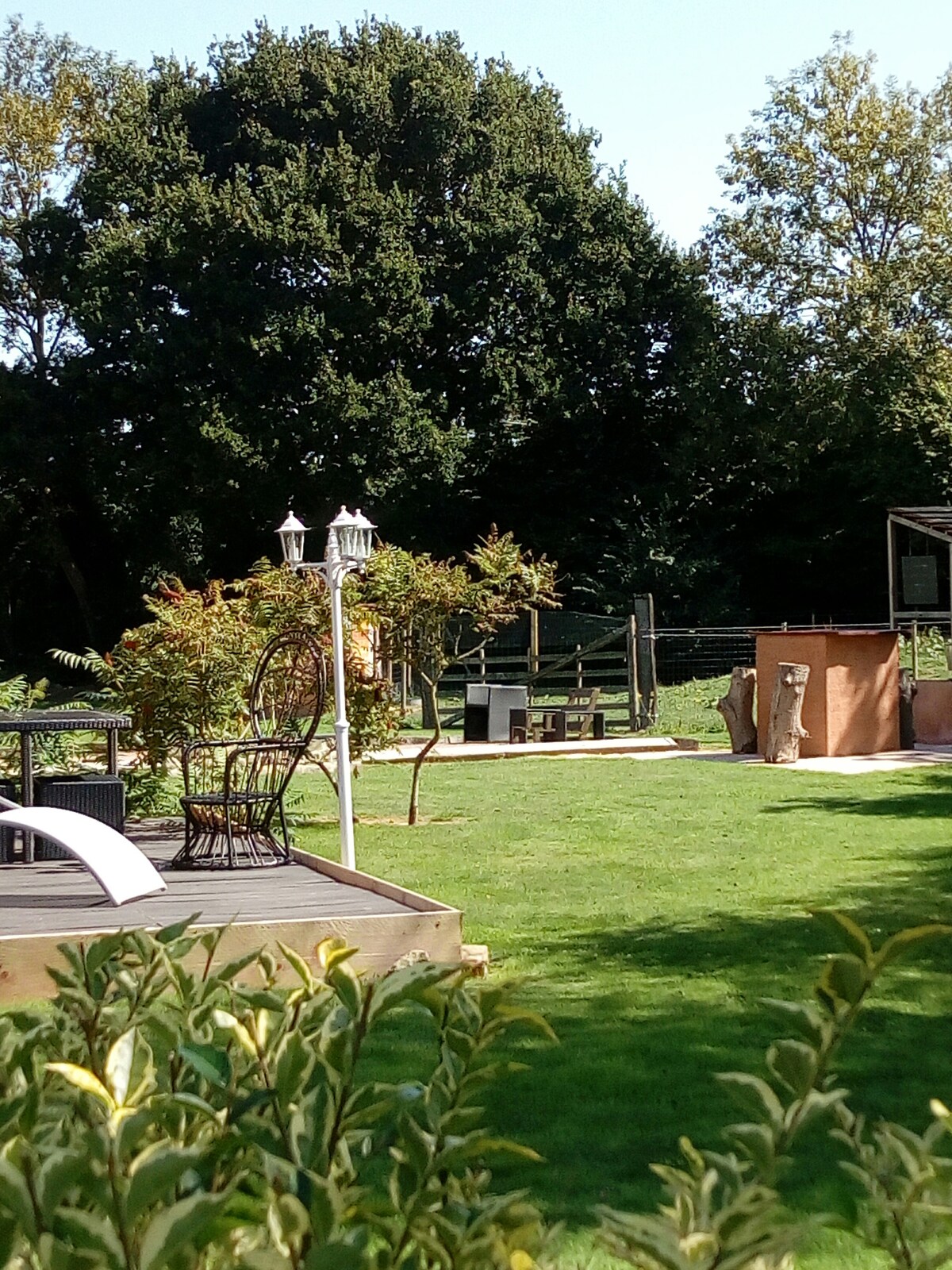 A serene outdoor space is visible, featuring a well-maintained lawn surrounded by trees. A wooden deck is highlighted with a lounge chair and a decorative lamp. In the background, a rustic structure and a wooden bridge can be seen, providing an inviting atmosphere.