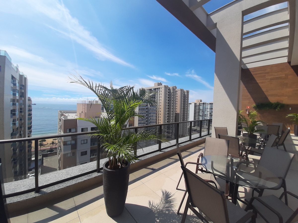 An outdoor balcony is presented, featuring a dining set for four and a potted palm. The view encompasses the ocean and surrounding high-rise buildings, with clear blue skies above. The area is well-lit, showcasing a spacious, inviting environment.