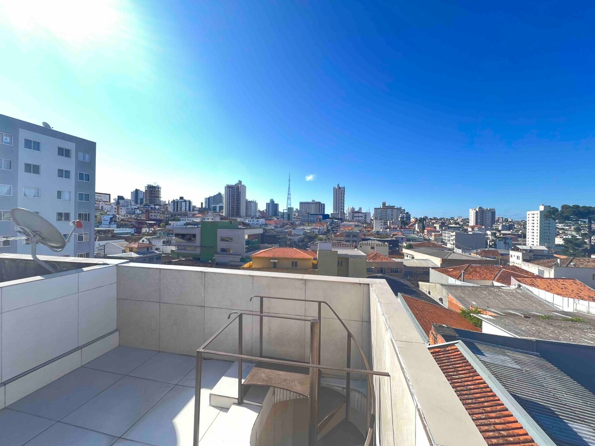 A rooftop terrace is visible, featuring a railing that frames a panoramic view of the city skyline. A spacious outdoor area is highlighted, with clear blue skies and scattered buildings in the distance emphasizing urban life.