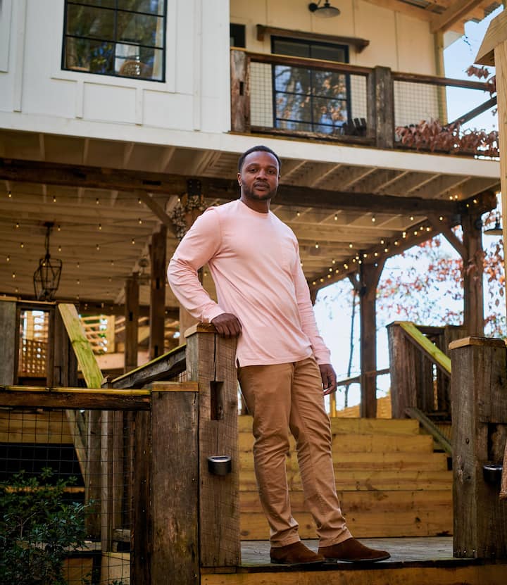 A man stands at the bottom of a short wooden staircase leading up to a raised house and covered patio.