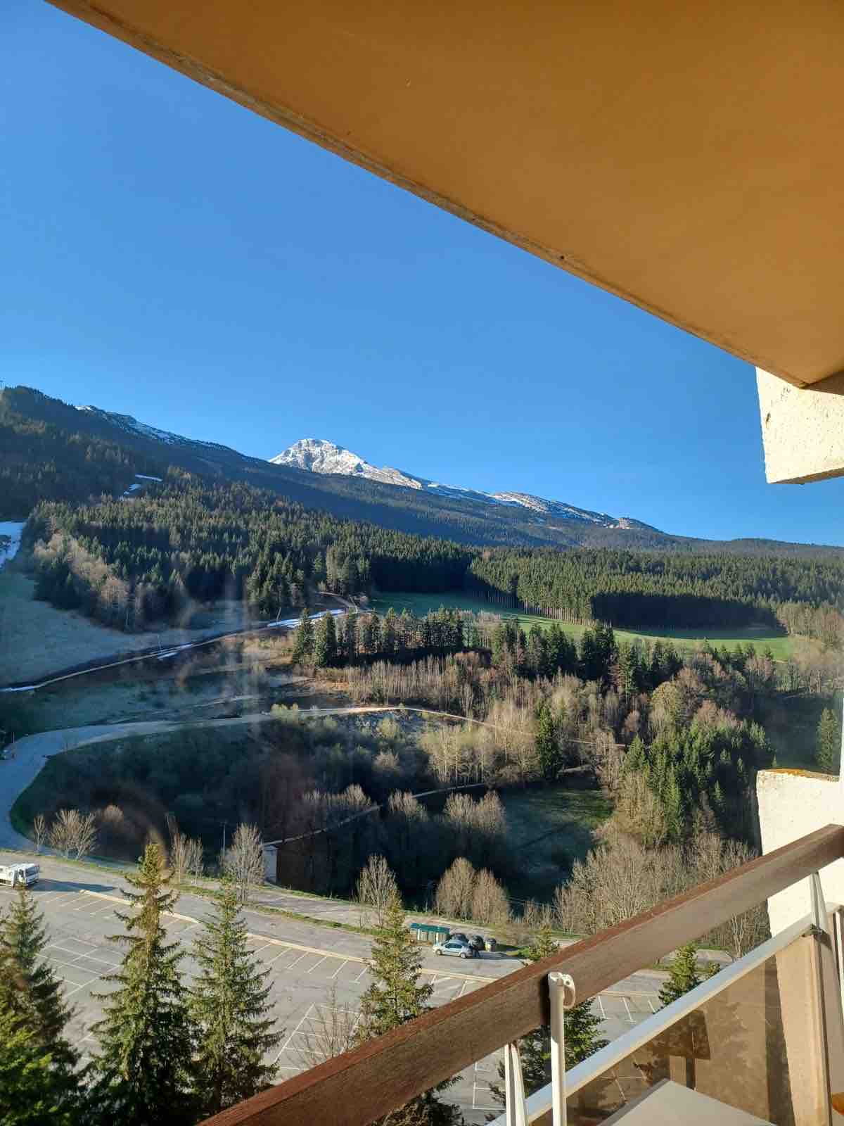 A scenic view is captured from the balcony, showcasing rolling green hills and mountain peaks in the background. The bright blue sky complements the landscape, creating a tranquil outdoor atmosphere. Trees line the lower area, enhancing the natural surroundings.