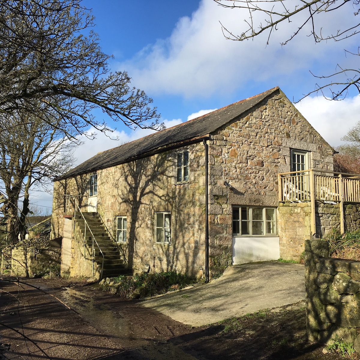 The exterior of a semi-detached barn is showcased, featuring textured granite stone walls and multiple large windows. A staircase leads to the entrance, while a balcony offers additional outdoor space. The setting reflects a peaceful rural environment with trees framing the structure.