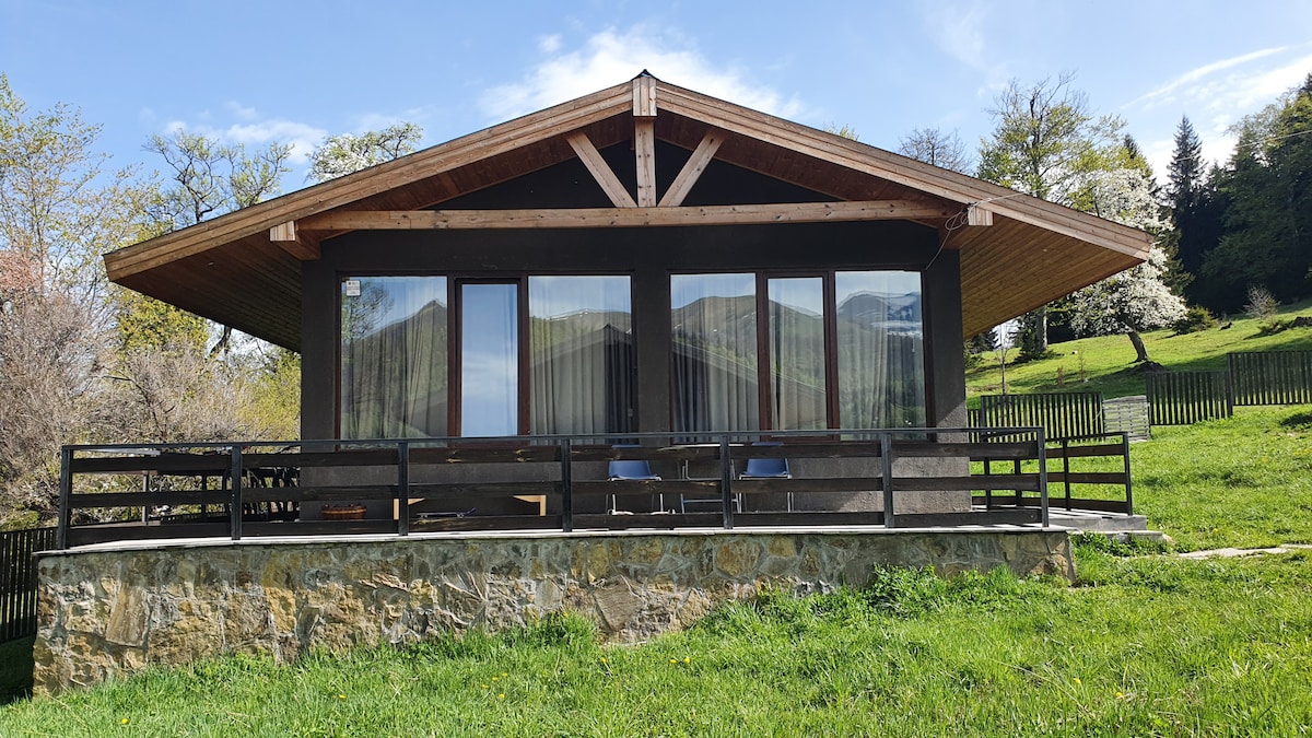The cottage features a spacious exterior with large glass windows framed by wooden beams. A broad terrace is lined with simple chairs, offering an unobstructed view of the surrounding greenery and the peaceful landscape.