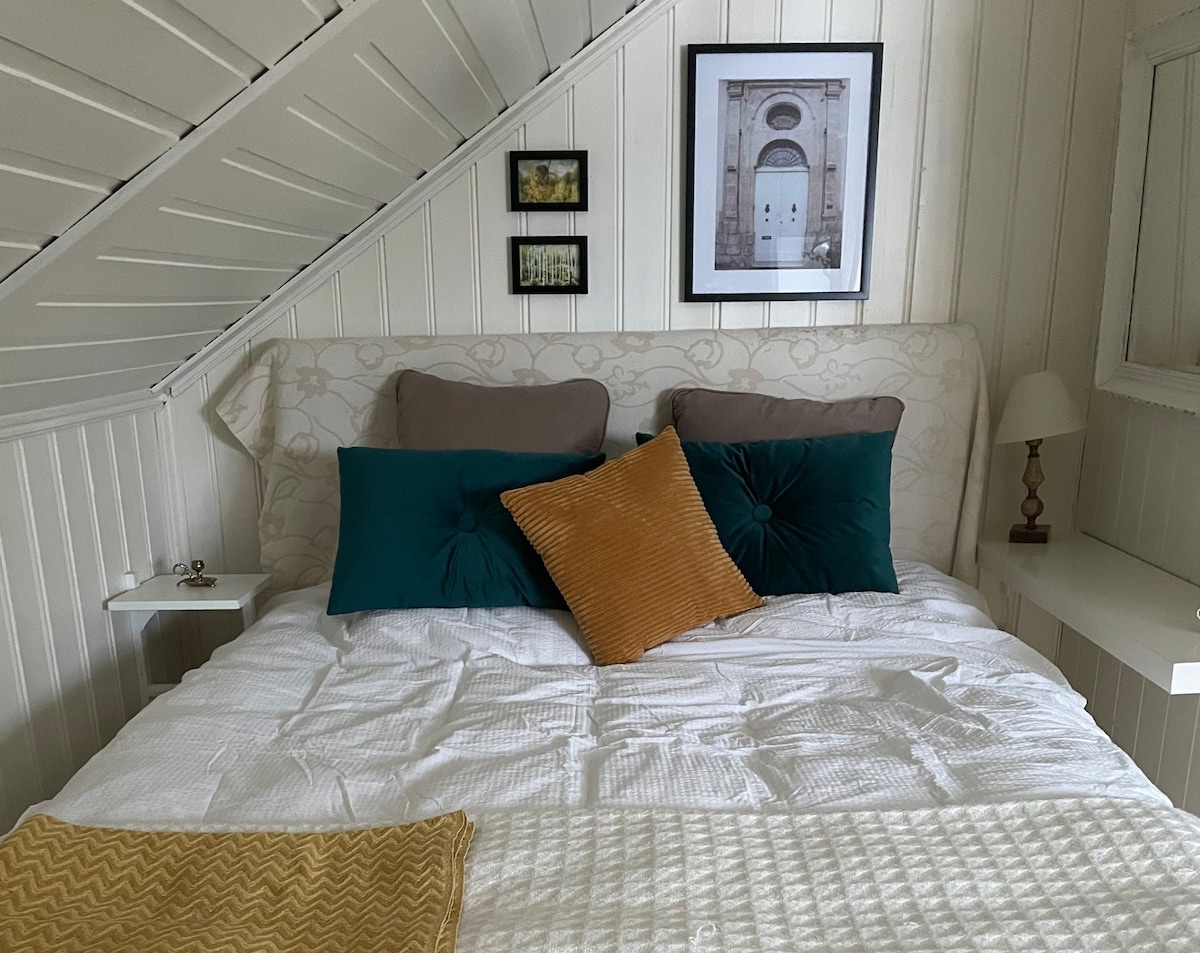 A cozy sleeping area features a spacious bed with soft linens and decorative pillows in teal and gold. Framed artwork hangs above the bed, enhancing the space. A bedside table with a lamp and a small shelf are positioned on one side, providing convenient surfaces.
