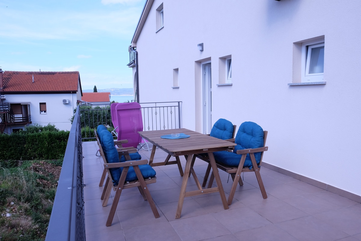 A spacious terrace is showcased, featuring a wooden table surrounded by six comfortable chairs with blue cushions. Natural light brightens the area, with a pink chair visible in the background. A view of the surrounding landscape is accessible from this outdoor space.