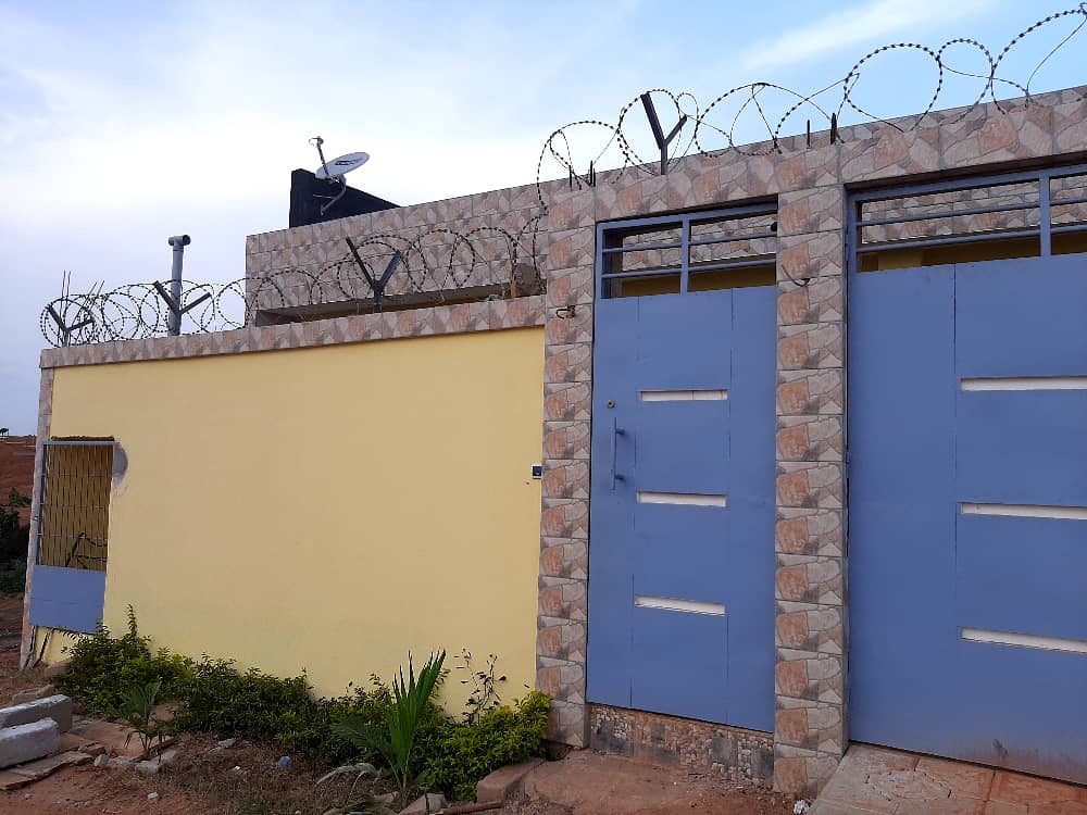 The exterior of the residence features a yellow wall complemented by stone accents and two blue doors. Barbed wire is visible atop the structure, along with a satellite dish. The surrounding area includes low greenery along the base of the wall.