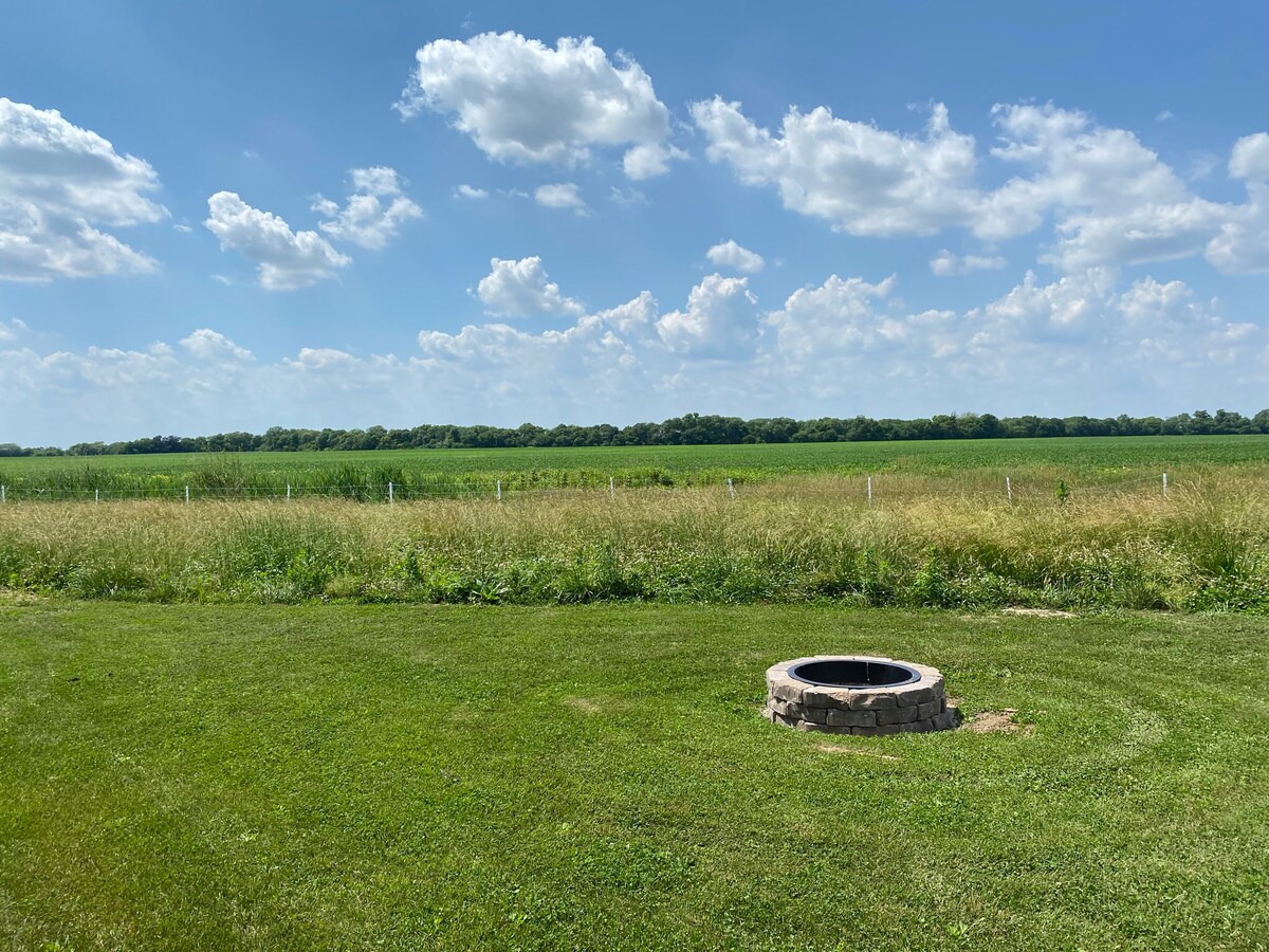 A spacious outdoor area features a circular fire pit surrounded by manicured grass. Lush green fields stretch into the distance under a bright blue sky adorned with fluffy white clouds.