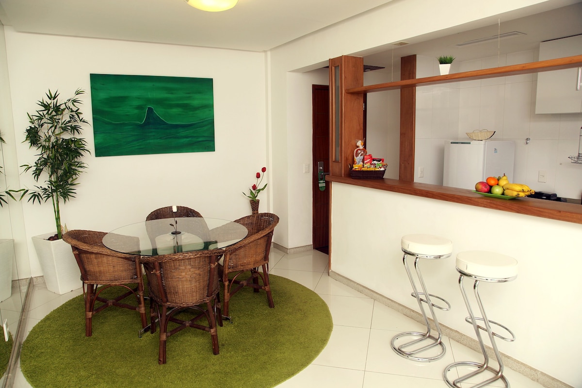 A modern dining area features a glass table surrounded by four wicker chairs on a plush green rug. A wooden bar area includes two high stools and fresh fruit displayed on the countertop. A painting of a wave adds a decorative touch to the wall.