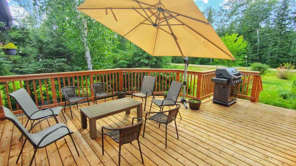 An outdoor deck is presented with light wooden flooring, featuring a large umbrella providing shade. Several chairs surround a central table, with a grill positioned to one side. Lush greenery and trees serve as a natural backdrop, enhancing the outdoor relaxation space.