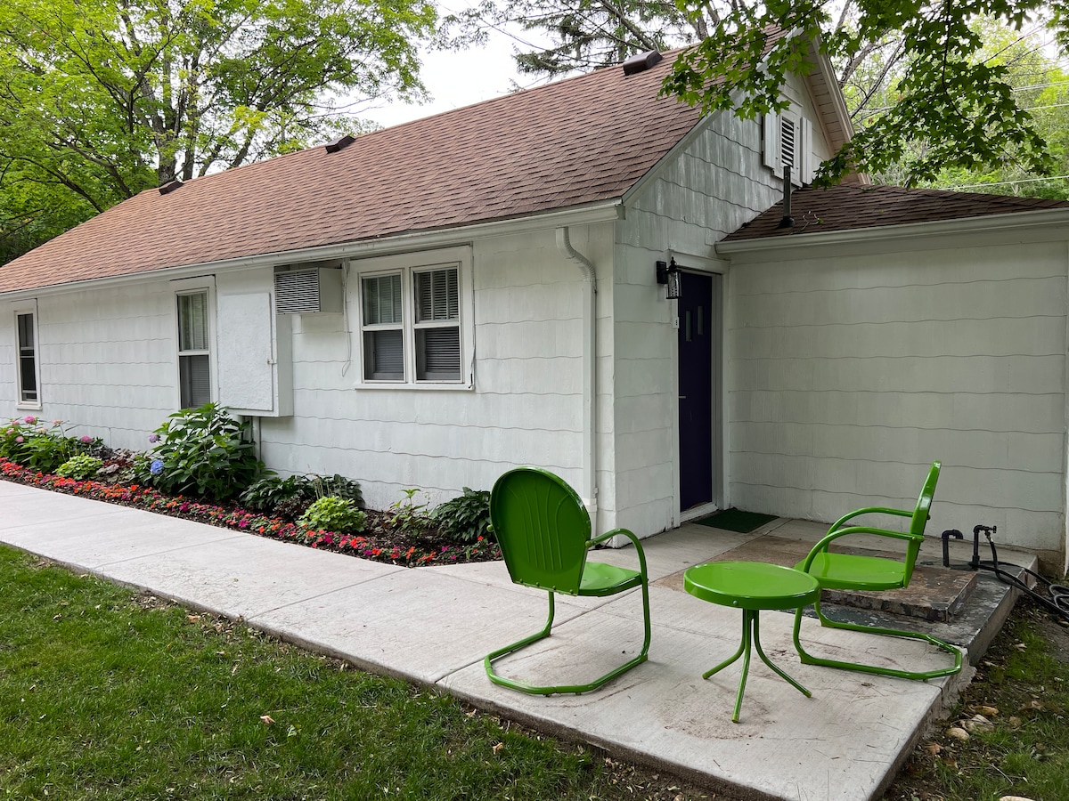 An outdoor area features a concrete path leading to a cozy seating arrangement. Two green chairs surround a small matching table, positioned beside flower beds filled with vibrant blooms. The cottage's white exterior is complemented by a brown roof and large windows.