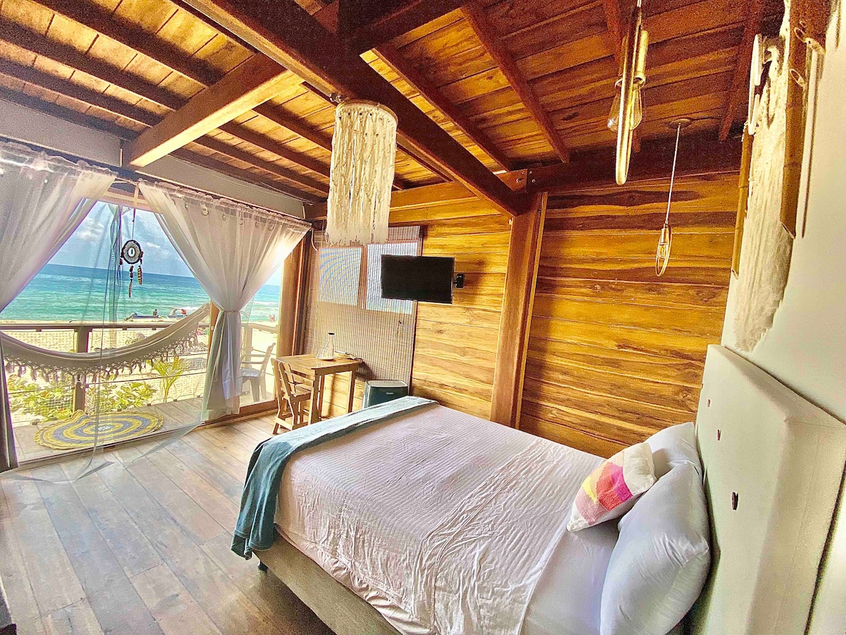 A spacious room features a comfortable bed with light bedding. Large windows provide a view of the ocean, with sheer curtains gently billowing. A wooden ceiling and wall add warmth, while a small table and chair are positioned nearby. A TV is mounted on the wall.