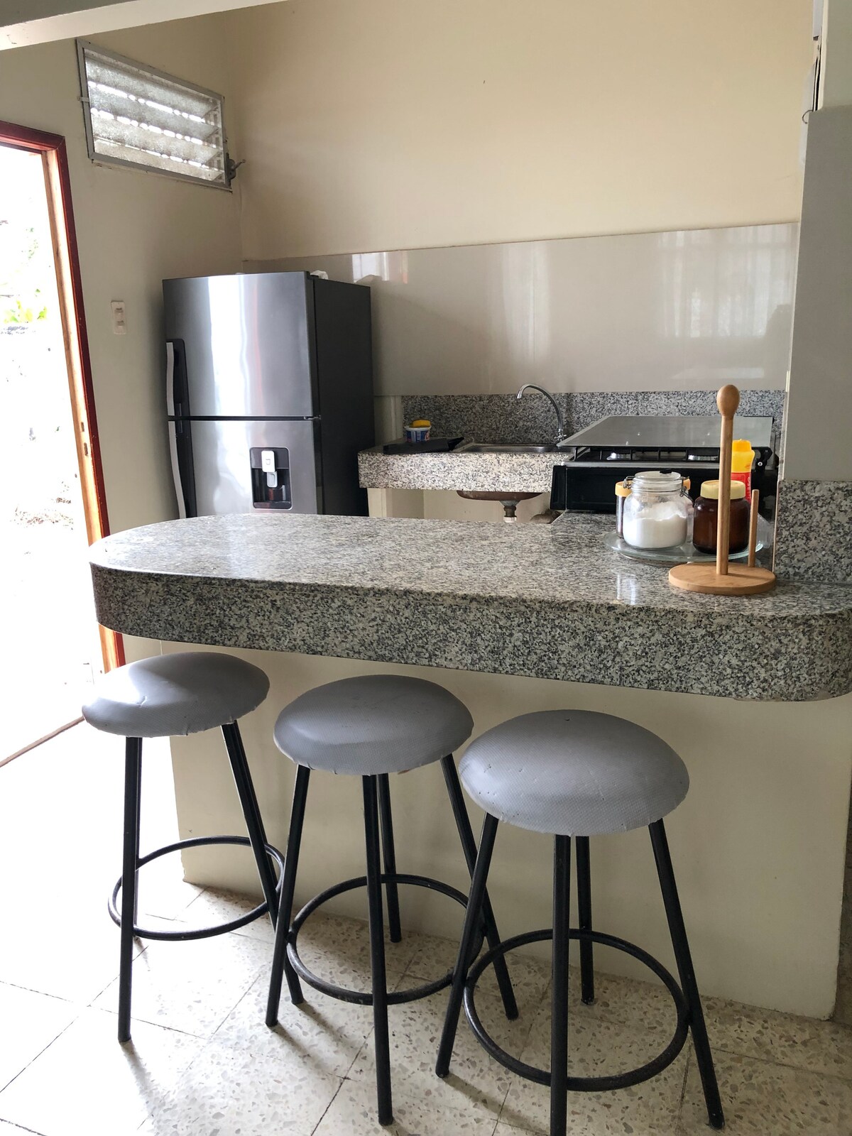 A compact kitchen area features a granite countertop with three gray bar stools. The modern refrigerator and stove are visible in the background, providing essential appliances for meal preparation. Natural light enters through an adjacent door, enhancing the space's functionality.