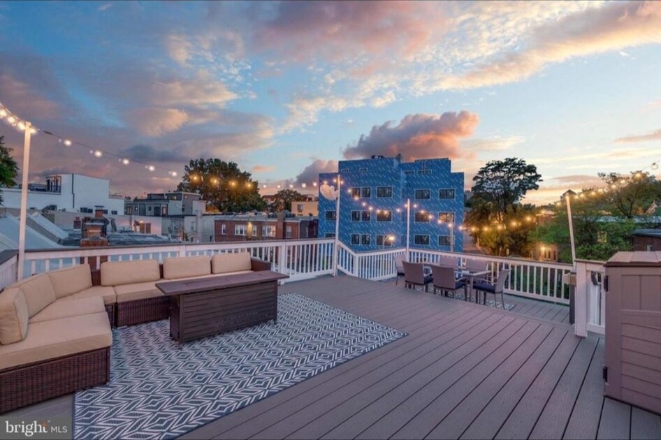 An expansive roof deck presents a cozy lounge area with sectional seating and a large coffee table. String lights are elegantly draped overhead, creating a warm ambiance as the sun sets behind the buildings in the distance.