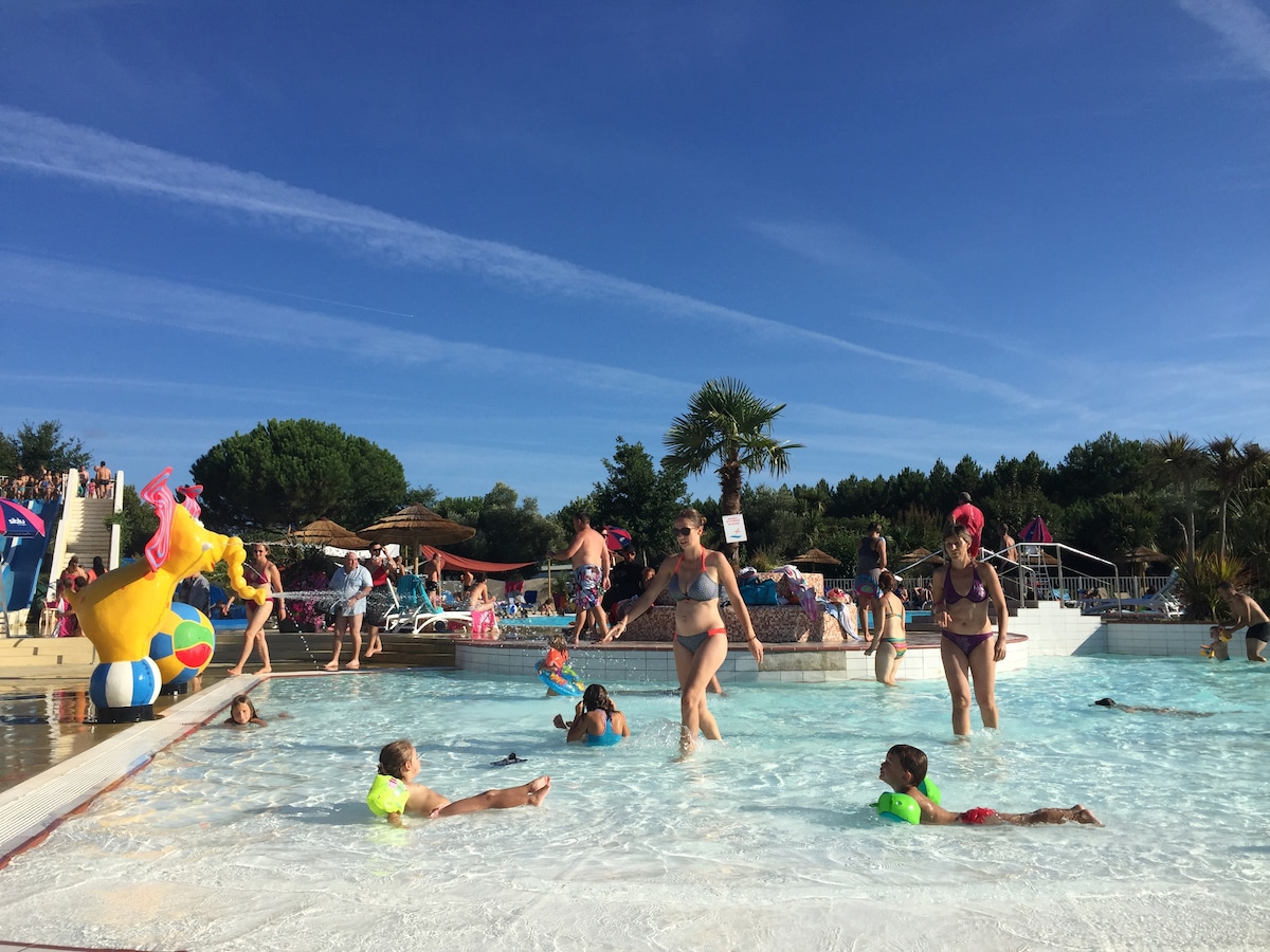 A spacious outdoor pool area is seen, featuring individuals of various ages enjoying the water. Colorful floats and beach balls are present, while lounge chairs and palm trees create a relaxed atmosphere. Clear blue skies complement the vibrant scene.