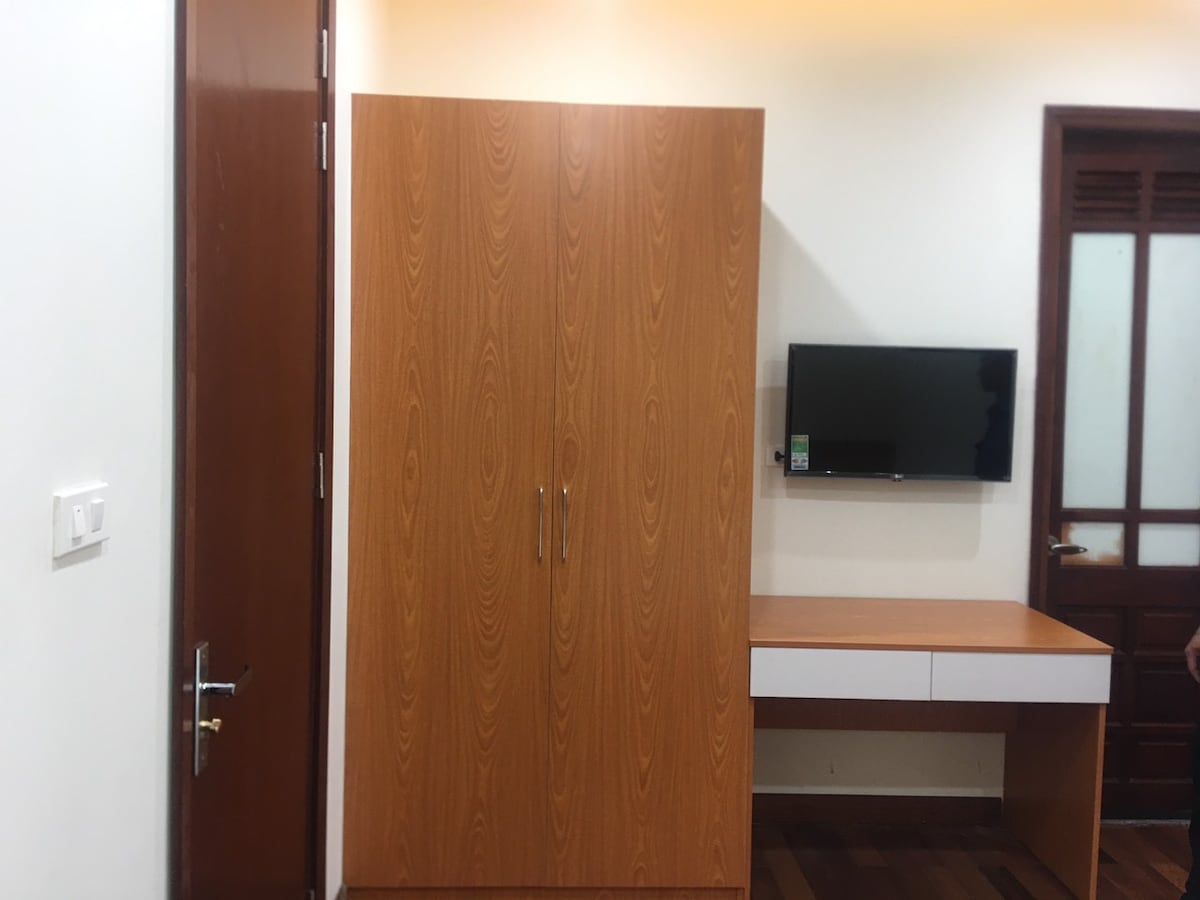 A wooden wardrobe stands against a wall, featuring smooth, minimalistic lines. Nearby, a small desk is positioned under a wall-mounted television, contributing to a functional workspace. A door with glass panels can be seen in the background, enhancing the room's overall brightness.