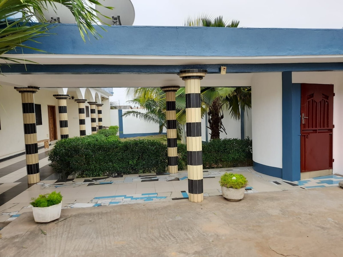 A covered walkway is depicted with distinctive column designs in black and white. Lush greenery is present on both sides, framed by ornamental plants in white pots. A red door adds a touch of color, leading into the inviting space beyond.