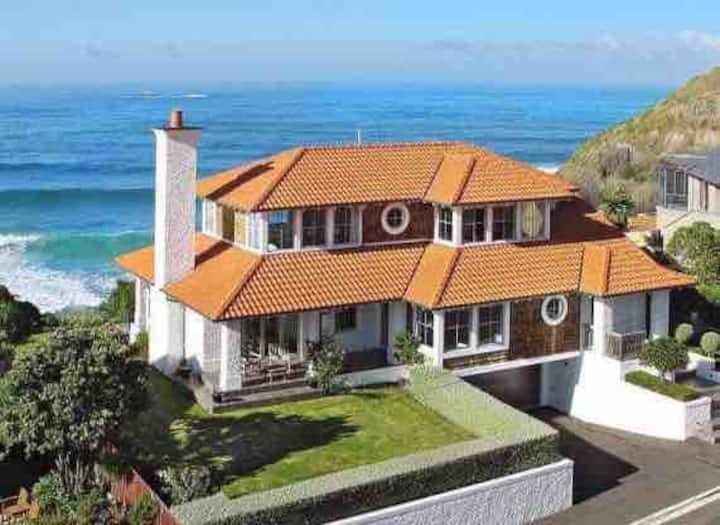 Elegant coastal home with a red-tiled roof, large chimney, and manicured garden, overlooking the ocean with clear blue skies