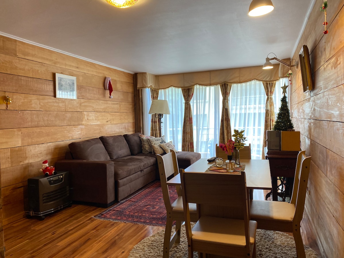 A living area is showcased with wooden walls, featuring a dark couch adorned with decorative pillows. A small dining table with two chairs is positioned nearby, while a window with sheer curtains allows natural light to brighten the space.