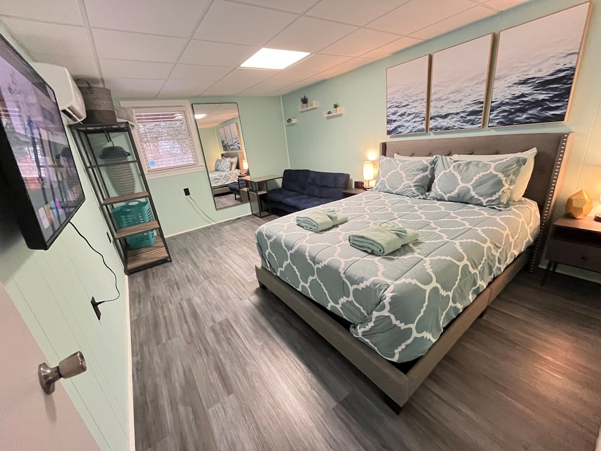 A newly remodeled bedroom features a queen-size bed adorned with soft linens and two folded towels. A futon couch is positioned nearby, and a smart TV is mounted on the wall. Natural light enters through a window, complementing the light blue walls and calming decor.