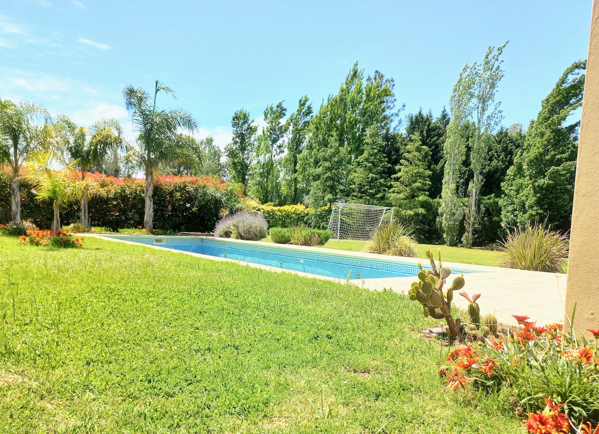 A serene swimming pool is set against a backdrop of lush greenery and colorful flowers. Palm trees line the space, while a soccer goal is visible in the distance, complementing the expansive lawn and garden area.