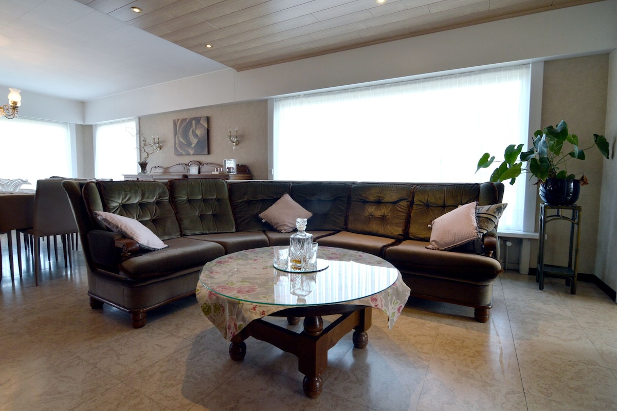 A comfortable living room is highlighted by a spacious leather sofa arranged in an L-shape. A round coffee table sits at the center, adorned with a decorative piece. Natural light pours in through large windows, offering a bright and inviting ambiance.