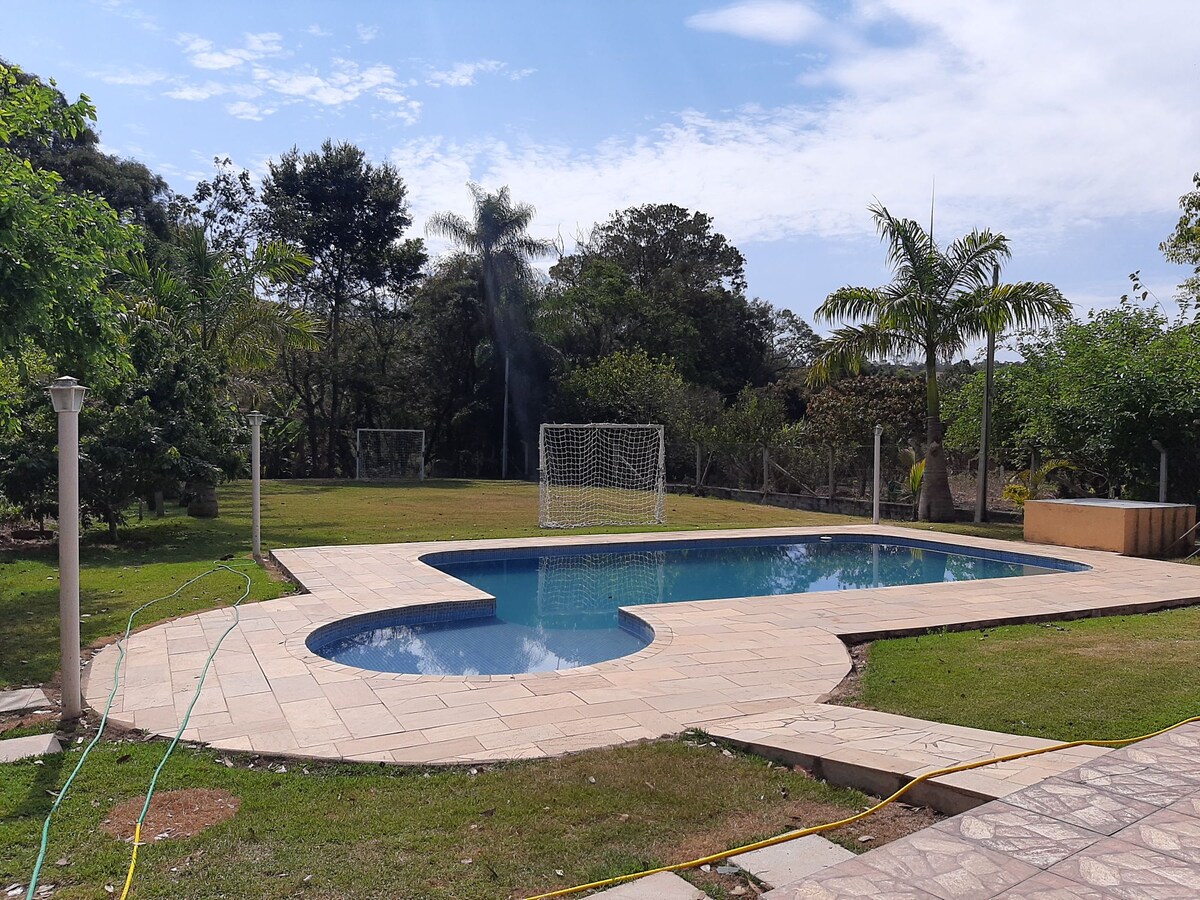 An outdoor pool, shaped like a pebble, is surrounded by a paved patio and lush green lawn. A soccer goal is visible in the background, framed by various trees. The clear sky and soft clouds enhance the serene setting.