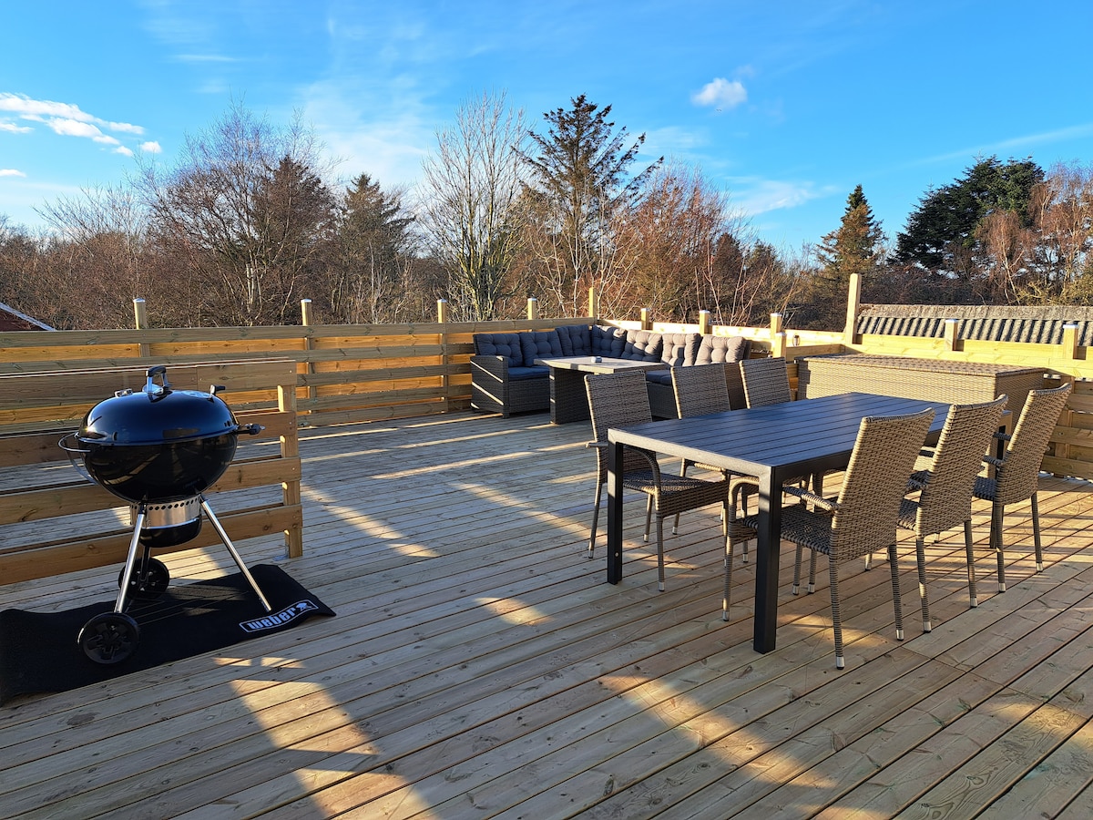 A spacious rooftop terrace is featured, furnished with a dining table and chairs, along with a seating area. A charcoal grill is positioned on a mat, complementing the wooden decking. Trees and clear blue skies form the backdrop, enhancing the outdoor experience.