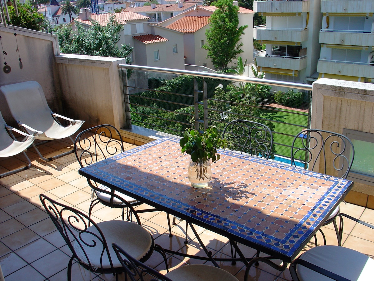 An outdoor terrace features a tile-topped table surrounded by six metal chairs. A potted plant is positioned at the center of the table. Views of green trees and nearby buildings are visible through the glass railing, offering a serene setting.