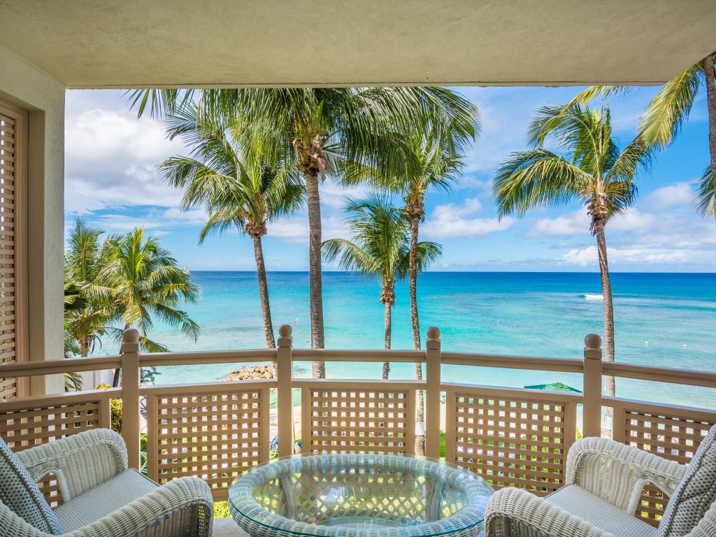 A serene terrace view reveals swaying palm trees against the backdrop of the sparkling Caribbean Sea. The glass table and woven chairs create a comfortable space to enjoy the coastal panorama, complemented by clear blue skies and gentle waves in the distance.