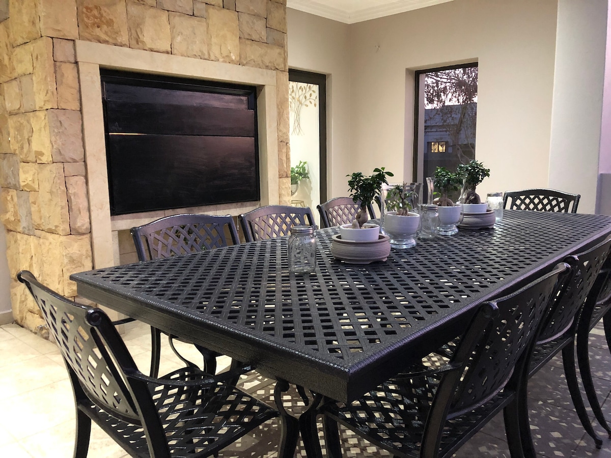 An outdoor dining area is featured with a large, dark metal table surrounded by matching chairs. Potted green plants are arranged on the table next to glassware and white dishware. A stone wall with a fireplace adds texture to the setting, while windows allow natural light to enter.