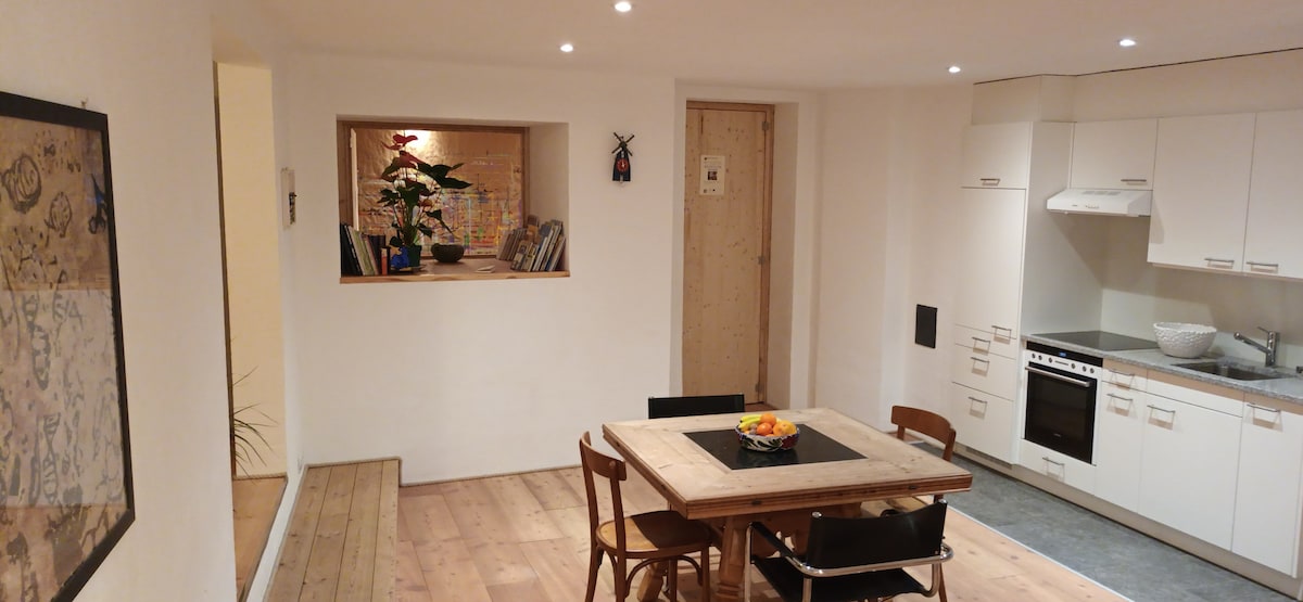 A bright and airy kitchen-dining space is observed featuring a wooden table surrounded by six chairs, complemented by natural light from recessed ceiling lights. The kitchen area includes a stove and a sink, with a small window providing a view of a potted plant.