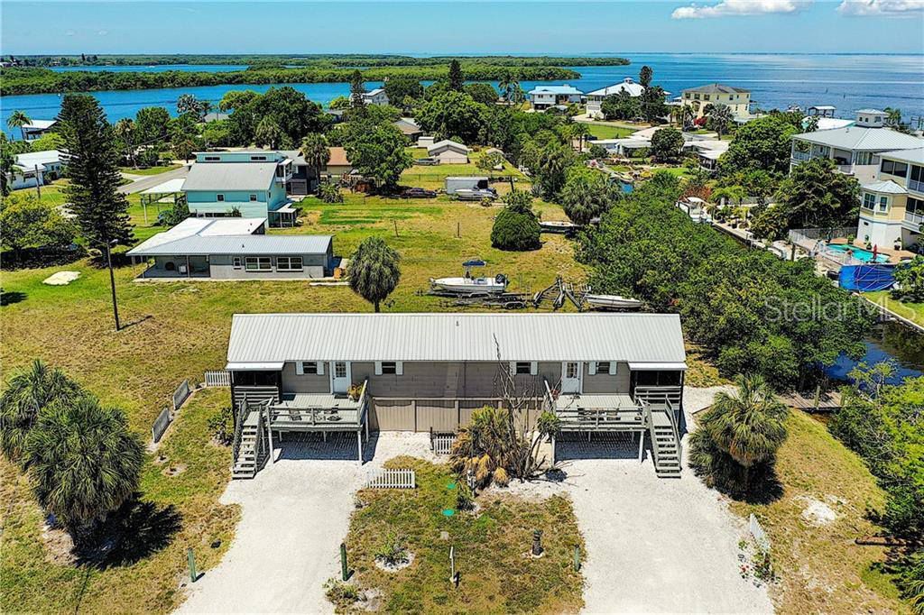 An aerial view of a duplex home, surrounded by greenery and featuring a private dock. Canal waters glisten in the distance, and neighboring homes are visible, adding to the serene atmosphere of the area. The structure is elevated on stilts, leading to a sandy pathway.