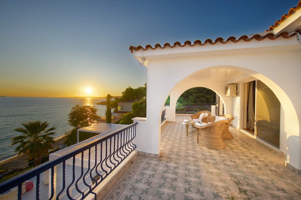 A spacious veranda features arched entryways and elegant tile flooring, offering a seating area with woven furniture. The sunset casts warm hues against the backdrop of the sea, while palm trees frame the view, enhancing the serene coastal atmosphere.
