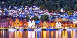A scenic view captures colorful waterfront buildings reflecting in calm waters, surrounded by lush green hills. The architectural styles vary from traditional to modern, with boats gently anchored along the shoreline, creating a welcoming ambiance in the evening light.