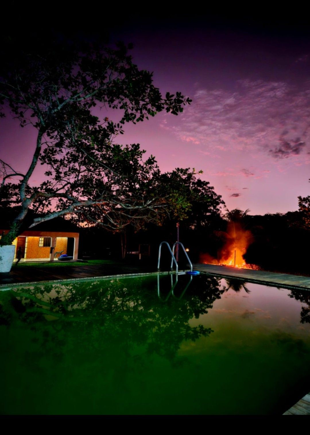 A tranquil scene features a pool reflecting a colorful twilight sky, with a cozy cabin visible in the background. A warm glow from a distant fire adds ambiance to the serene outdoor space surrounded by lush greenery.