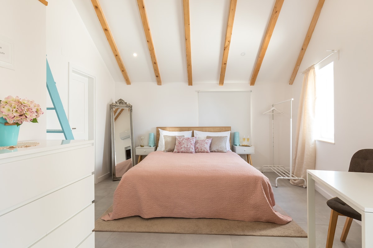 A spacious studio apartment features a large bed dressed in soft pink linens against a backdrop of natural wood beams. Light walls and a large window brighten the room, complemented by a mirror and a small workspace with a chair, creating an open and airy atmosphere.
