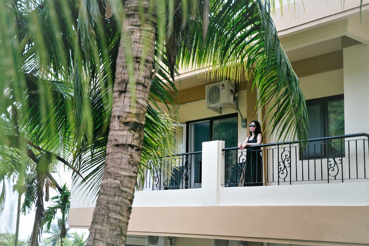 A balcony is visible, adorned with black railings and surrounded by lush palm trees. Large windows behind the balcony provide a view into the villa. A person stands near the edge, blending into the tranquil outdoor setting.