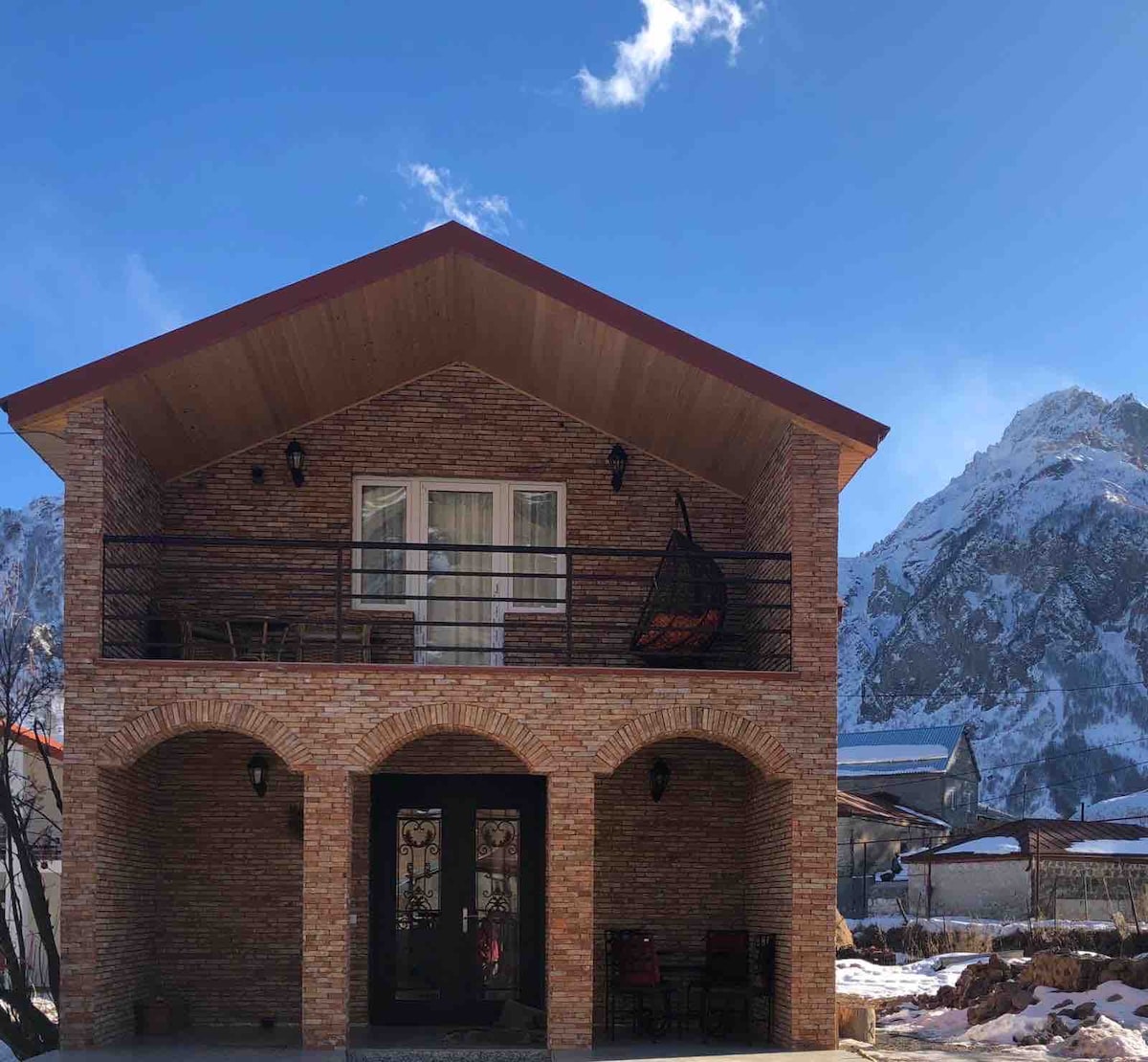 A two-story brick house is depicted, featuring a sloped roof and a spacious front porch with arched entrances. A balcony is visible on the second floor, with seating arrangements that overlook the mountain scenery. Clear blue skies and mountain peaks create a stunning backdrop.