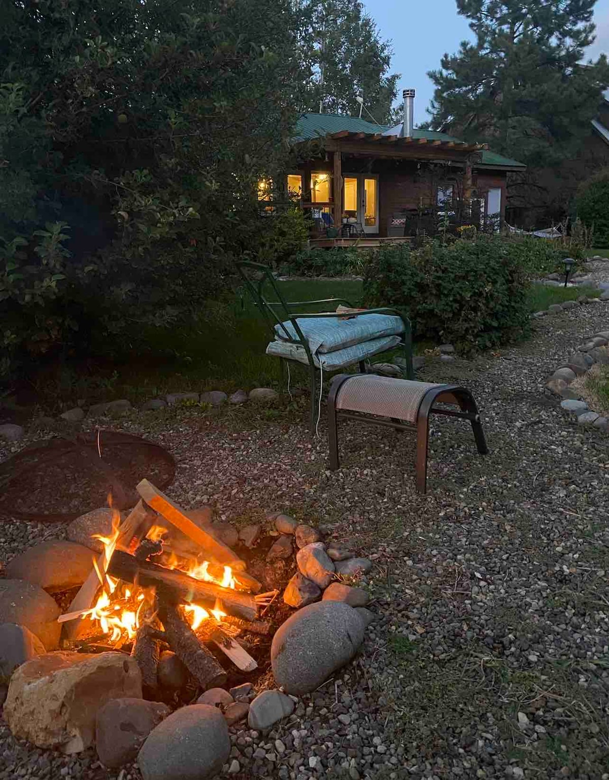 A cozy fire pit is surrounded by smooth stones, with a seated area featuring a metal chair and a small table nearby. The lush garden and trees create a secluded atmosphere in the evening light, with a home visible in the background.