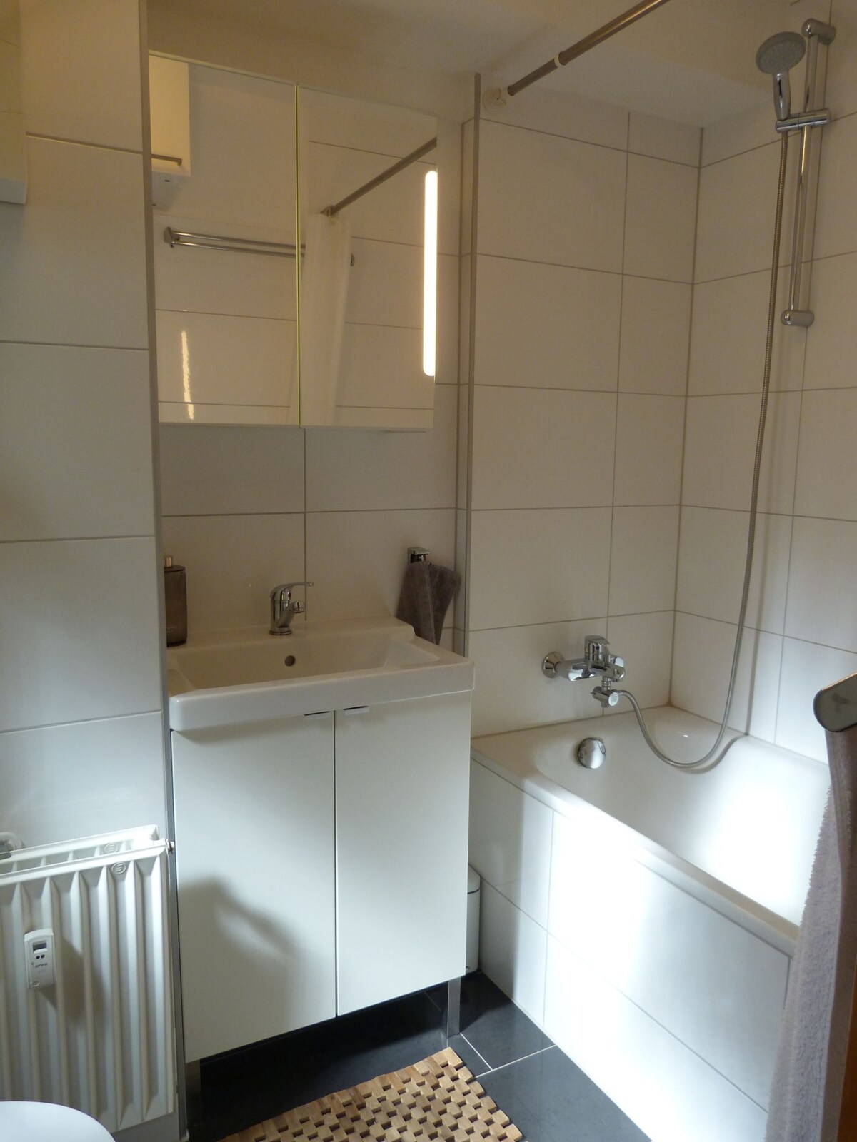A modern bathroom is shown, featuring a clean, white tiled design. A bathtub is positioned alongside a compact sink and mirrored cabinet. Light reflects off the shiny surfaces, while a towel rack holds a grey towel. A small rug is placed on the dark tiled floor.