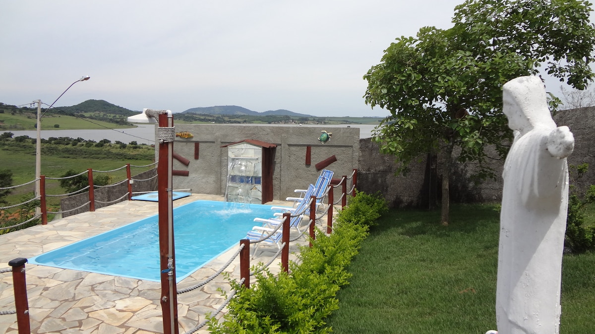 A swimming pool is bordered by stone tiles and surrounded by green grass. Lounge chairs are positioned around the pool area. A statue stands nearby, overlooking the landscape, which features views of the lake and distant hills.