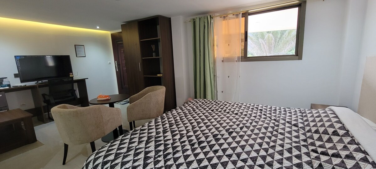 A spacious guest room features a large bed with a geometric patterned blanket. Natural light filters through a window adorned with sheer curtains, illuminating a work desk and two upholstered chairs. A TV is mounted on a dark wood cabinet, adding functionality to the space.