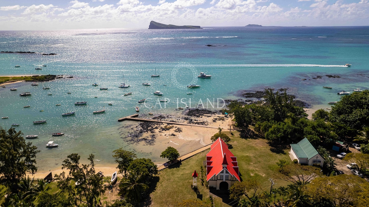 Aerial view of a scenic coastal area featuring a sandy beach and clear blue waters. The image showcases a red-roofed building surrounded by greenery, with several boats anchored nearby and distant islands visible on the horizon.