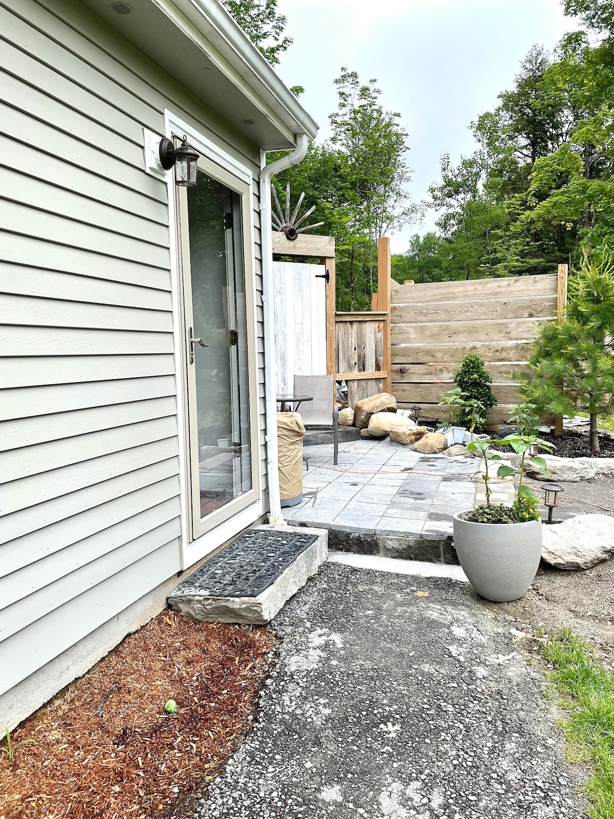 The exterior of the guest suite is shown, featuring a stone pathway leading to a glass door. Landscaping includes a decorative planter and natural stone elements, surrounded by greenery. A wooden fence provides privacy, creating a welcoming entryway to the space.