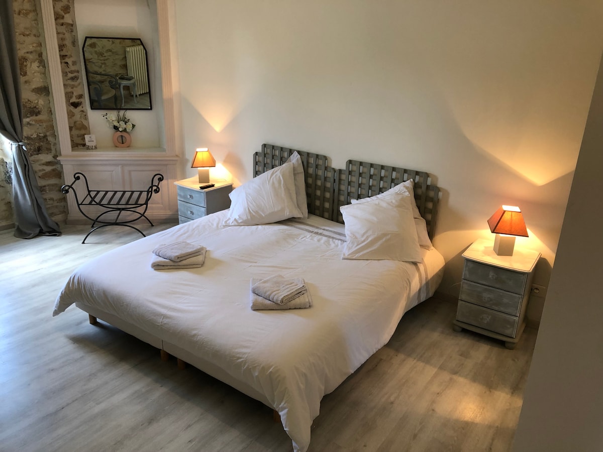 A spacious sleeping area features a large bed with soft linens and two decorative pillows. Bedside tables with bedside lamps provide ambient lighting, while a subtle wall mirror enhances the bright atmosphere. A comfortable chair completes the inviting layout of the room.