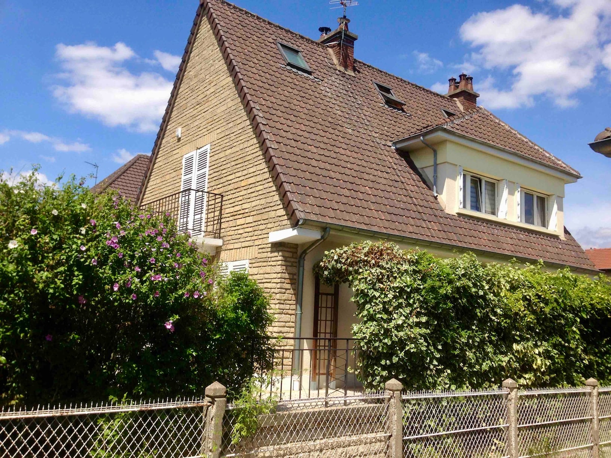 A two-story house is nestled amidst lush greenery and flowering plants. The structure features a sloped roof with skylights and multiple windows. A small balcony is visible on the upper floor, while a low fence borders the property, enhancing the private outdoor space.