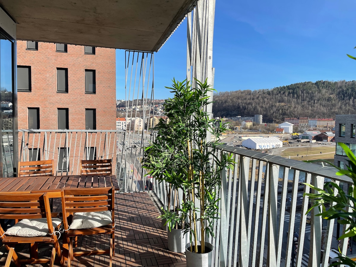 A spacious balcony features a wooden dining table surrounded by matching chairs. Tall planters with greenery provide a touch of nature. The view extends to distant hills and the city, with sunlight shining brightly against a clear blue sky.